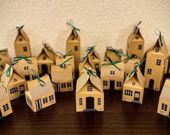 May include: A brown paper advent calendar with 24 small houses, each with a different number. The houses are decorated with green ribbon and have small windows. The calendar is sitting on a wooden surface.