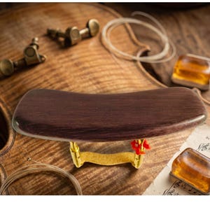 May include: A dark brown violin shoulder rest with a curved wooden pad and gold-coloured metal supports. The rest is shown on a wooden surface with other violin accessories, including tuning pegs, strings, and rosin.