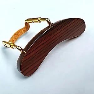 May include: A violin shoulder rest featuring a curved, dark brown wooden pad with a wood grain pattern and gold-coloured metal hardware. A tan-coloured, textured cushion is attached to the metal hardware.