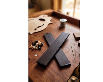 May include: Two rectangular, dark-toned wooden fingerboard blanks arranged in an X shape. Nearby are small wood shavings, a spool of silver wire, two thin metal rods, a light-colored wooden instrument body part, and a metal hand plane tool.