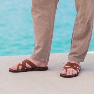 May include: A pair of brown leather sandals with a criss-cross strap design. The sandals are worn by a person standing on a concrete surface near a body of water.