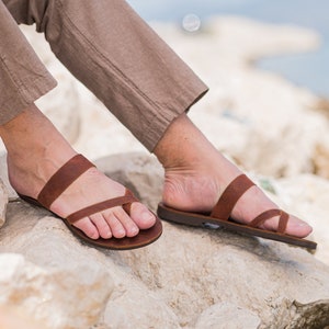 Pu&ograve; includere: Un paio di sandali in pelle marrone con due cinturini sul piede. Le sandali sono indossate da una persona seduta su rocce.
