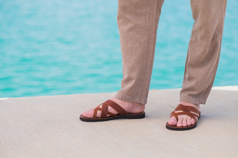 May include: A pair of brown leather sandals with a criss-cross strap design. The sandals are worn by a person standing on a concrete surface near a body of water.
