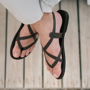May include: A pair of black leather sandals with criss-cross straps. The sandals are worn on a person's feet.