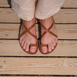 May include: A pair of brown leather sandals with criss-cross straps and a toe loop. The sandals are worn on a wooden deck.