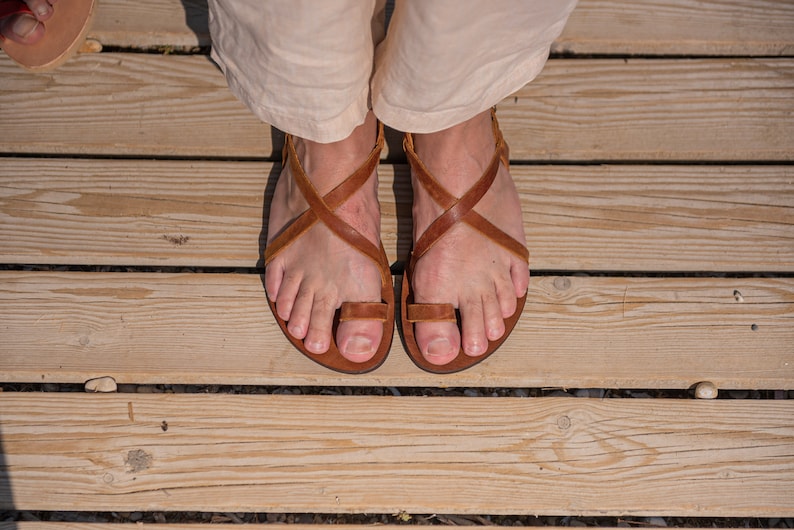 May include: A pair of brown leather sandals with criss-cross straps and a toe loop. The sandals are worn on a wooden deck.