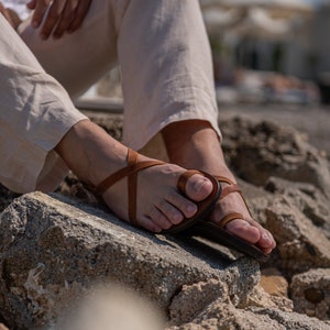 May include: A pair of brown leather sandals with a criss-cross strap design. The sandals are being worn on someone's feet, resting on a rocky surface.