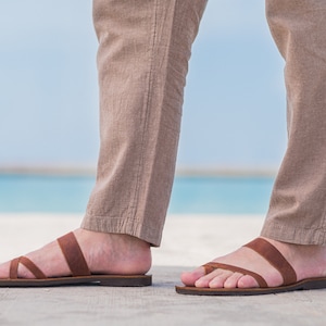 Pu&ograve; includere: Un paio di sandali in pelle marrone con due cinturini sul piede. Le sandali sono indossate da una persona in piedi su una superficie di cemento con una spiaggia e l'oceano sullo sfondo.