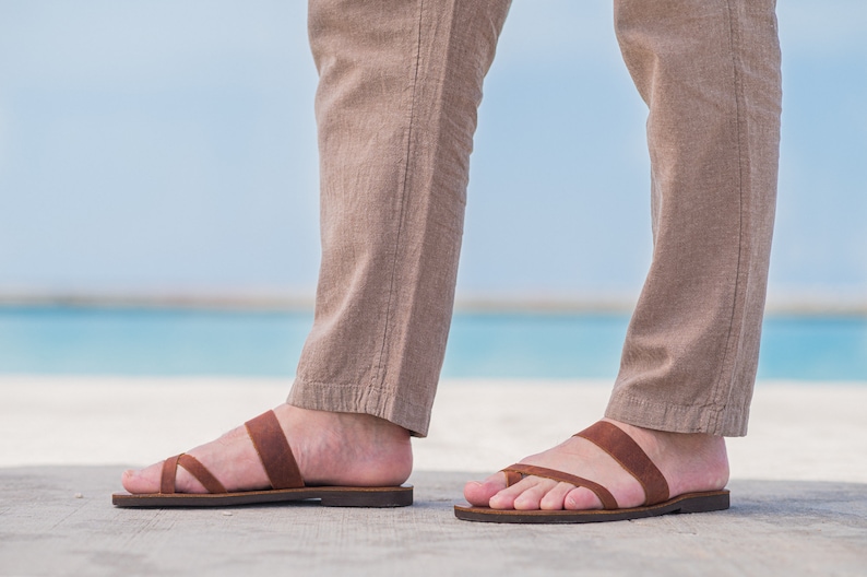 Pu&ograve; includere: Un paio di sandali in pelle marrone con due cinturini sul piede. Le sandali sono indossate da una persona in piedi su una superficie di cemento con una spiaggia e l'oceano sullo sfondo.
