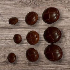 May include: Nine brown leather buttons with a stitched design. The buttons are arranged in three rows on a wooden surface.