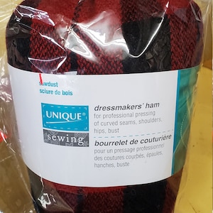 May include: A dressmakers' ham, packaged in plastic, featuring a red and black plaid design. The label includes the text "UNIQUE sewing" and text in English and French, detailing its use for pressing curved seams, shoulders, hips, and bust.