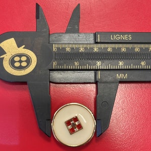 Peut inclure: Un bouton blanc avec un bord dor&eacute; et un motif &agrave; carreaux rouge et blanc au centre. Le bouton est mesur&eacute; avec une r&egrave;gle qui indique "LIGNES" et "MM".