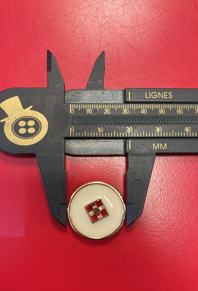 Peut inclure: Un bouton blanc avec un bord dor&eacute; et un motif &agrave; carreaux rouge et blanc au centre. Le bouton est mesur&eacute; avec une r&egrave;gle qui indique "LIGNES" et "MM".