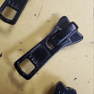 May include: Three black zipper pulls. The top pull has the text "YKK" on it. The middle pull has the text "YKK" on it. The bottom pull has the text "YKK" on it.