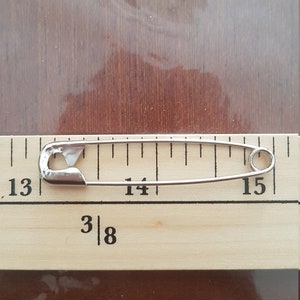 May include: A silver safety pin measuring 1 3/8 inches long, placed on a wooden ruler with markings from 13 to 15.