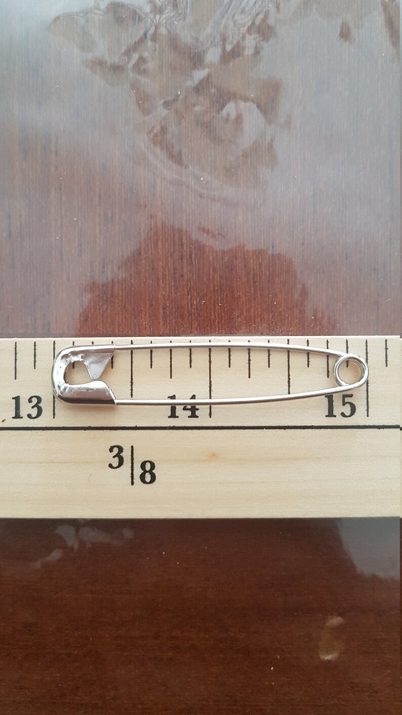 May include: A silver safety pin measuring 1 3/8 inches long, placed on a wooden ruler with markings from 13 to 15.