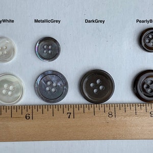 May include: Six different coloured buttons with four holes each. The buttons are arranged in two rows of three. The buttons are on a white surface with a wooden ruler underneath. The ruler is marked with inches. The buttons are pearly white, metallic grey, dark grey, pearly brown, pearly white, and metallic grey.