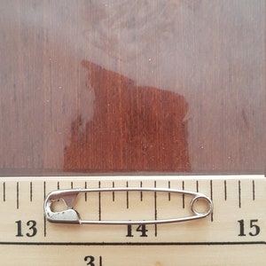 May include: A silver safety pin measuring 1 3/8 inches long, placed on a wooden ruler with markings from 13 to 15.