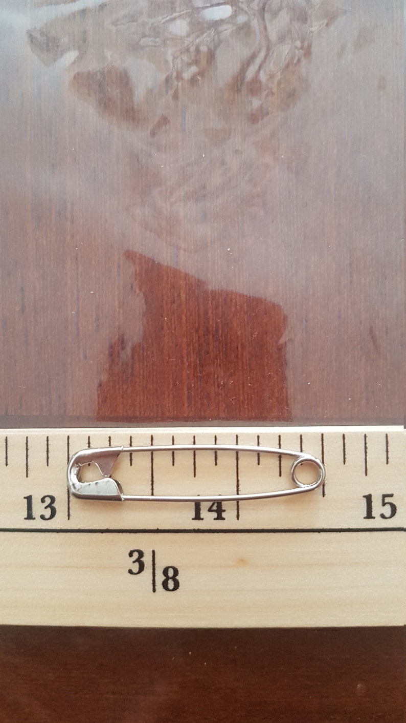May include: A silver safety pin measuring 1 3/8 inches long, placed on a wooden ruler with markings from 13 to 15.