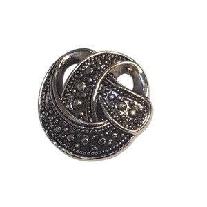 Peut inclure: Un bouton argenté avec un motif de nœud. Le bouton a une surface texturée avec de petits points en relief. Le nœud est formé par des boucles entrelacées, créant un élément décoratif. Le bouton est probablement destiné aux vêtements ou à l'artisanat.