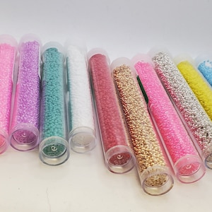 May include: Assortment of small glass seed beads in various colours, including orange, black, pink, purple, green, white, red, gold, silver, light pink, yellow, blue, and light orange, displayed in clear plastic tubes.
