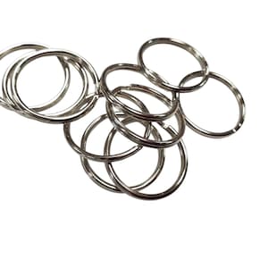 May include: A collection of shiny, silver-colored split key rings. The rings are made of metal and are arranged in a loose cluster against a white background. The key rings are round and have a split design for easy attachment.
