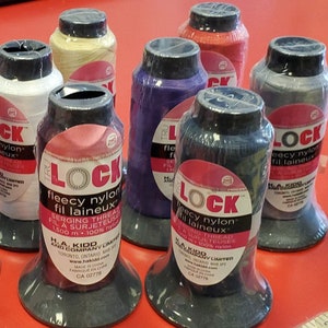 May include: Assortment of colourful spools of Lock brand serging thread. The spools are labelled "fleecy nylon fil laineux" and "serging thread fil a surjeteuse".