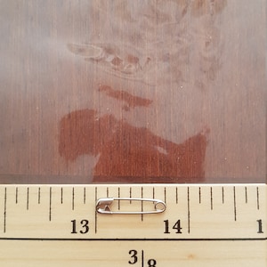 May include: A silver safety pin measuring approximately 1 3/8 inches in length, positioned on a wooden ruler with markings for inches and fractions of an inch.