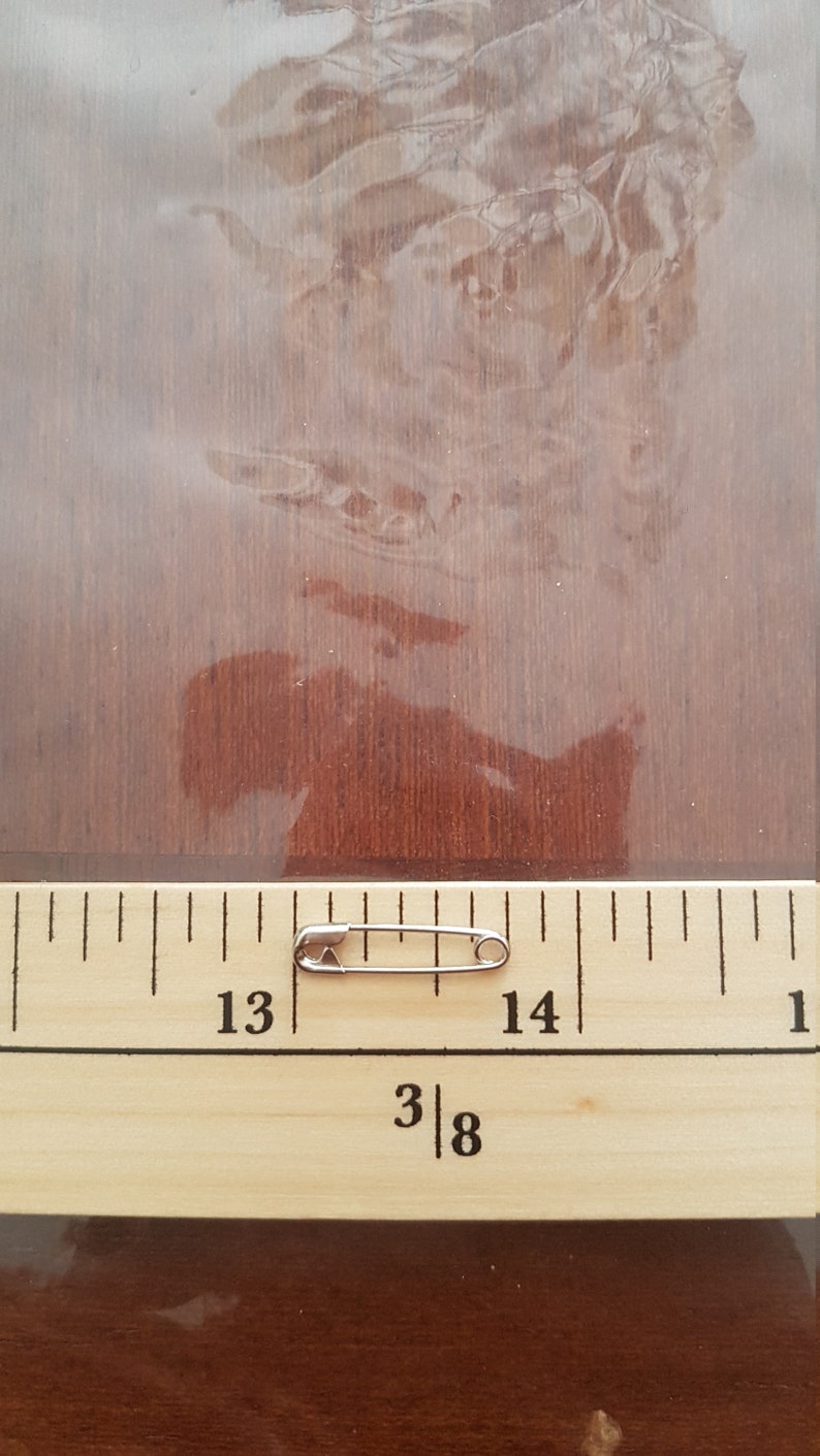 May include: A silver safety pin measuring approximately 1 3/8 inches in length, positioned on a wooden ruler with markings for inches and fractions of an inch.