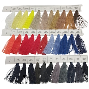 May include: A color chart featuring three rows of numbered thread samples. Each row displays various shades of embroidery floss arranged in a gradient, including white, yellow, tan, brown, orange, red, blue, grey, and black, with labels.
