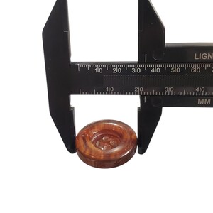 May include: A black caliper measuring a round, brown button. The button has a four-hole design and a wood-grain appearance. The caliper is marked in millimetres.