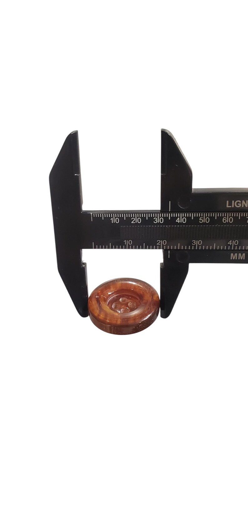 May include: A black caliper measuring a round, brown button. The button has a four-hole design and a wood-grain appearance. The caliper is marked in millimetres.