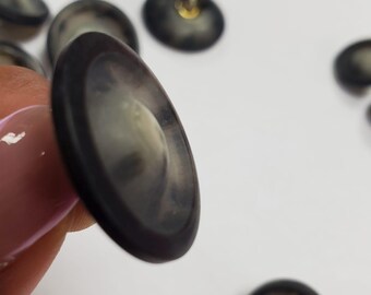 Peut inclure: Une collection de boutons ronds gris foncé avec un aspect translucide et marbré. Chaque bouton présente une surface lisse légèrement bombée et un bord extérieur sombre. Un bouton est tenu au premier plan, montrant son profil et sa profondeur.