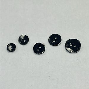 May include: A collection of five black buttons with two holes each. The buttons vary in size, with a glossy finish. The buttons are arranged on a white surface.