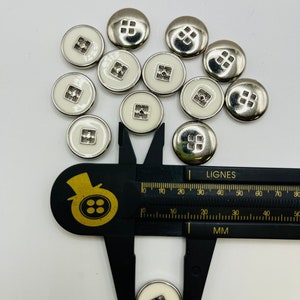 Peut inclure: Gros plan sur un groupe de boutons blancs avec une bordure argent&eacute;e et un motif carr&eacute; au centre. Les boutons sont mesur&eacute;s avec une r&egrave;gle, montrant qu'ils mesurent environ 1,5 cm de diam&egrave;tre.