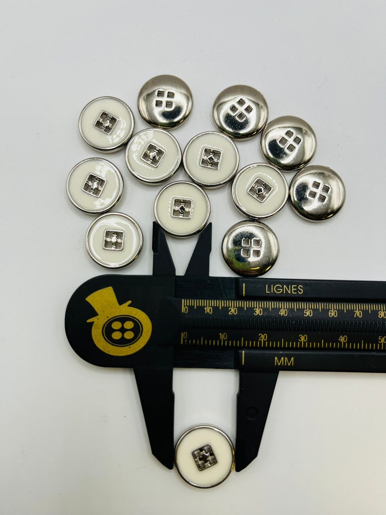 Peut inclure: Gros plan sur un groupe de boutons blancs avec une bordure argent&eacute;e et un motif carr&eacute; au centre. Les boutons sont mesur&eacute;s avec une r&egrave;gle, montrant qu'ils mesurent environ 1,5 cm de diam&egrave;tre.