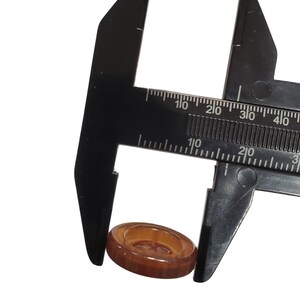 May include: A brown, oval button is measured with a black caliper. The button is made of a translucent material. The caliper is marked with measurements in centimetres and millimetres.