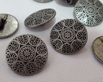 Könnte beinhalten: Eine Ansammlung runder, silberfarbener Metallknöpfe mit einem detaillierten, erhabenen geometrischen Muster und einem zentralen Blumenmotiv. Jeder Knopf hat eine glatte, leicht verwitterte Oberfläche und eine Öse auf der Rückseite.
