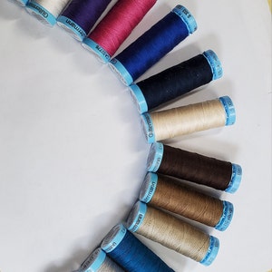 May include: A collection of 14 spools of thread in various colours, including black, white, purple, pink, blue, brown, beige, and teal. The spools are arranged in a semi-circle on a white surface. Each spool has a blue label with the brand name "Gütermann" and the text "100m-110 yds/vgs".