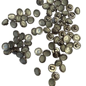 May include: A scattering of silver-coloured buttons with a pearlescent, oval-shaped centre. The buttons are spread across a white background, highlighting their shiny, metallic rims and the subtle iridescence of the centres. These buttons are suitable for sewing and craft projects.