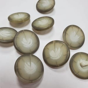Peut inclure: Un ensemble de boutons ronds et translucides présentant un design dégradé passant d'un bord vert olive foncé à un centre plus clair et crémeux. Chaque bouton affiche un motif blanc en forme de feuille rayonnant du milieu vers le bord.
