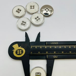 Peut inclure: Six boutons blancs avec une bordure argent&eacute;e et un motif carr&eacute; au centre. Les boutons mesurent 3,8 cm de diam&egrave;tre.