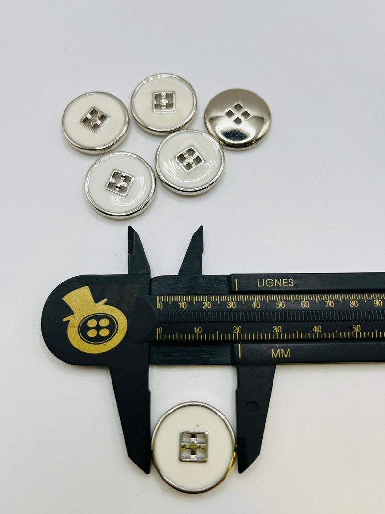 Peut inclure: Six boutons blancs avec une bordure argent&eacute;e et un motif carr&eacute; au centre. Les boutons mesurent 3,8 cm de diam&egrave;tre.