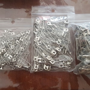 May include: Four bags of safety pins in various sizes. The bags are clear plastic and the pins are silver.