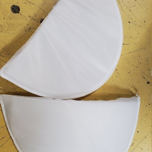 May include: Two white fabric bra inserts, shaped like half circles, with a sewn edge. The inserts are on a yellow surface.