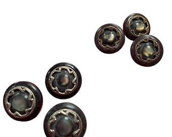 Könnte beinhalten: Ein Set aus fünf runden Knöpfen mit einem dunkelbraunen Außenrand und einem zentralen, schillernden grauen Cabochon. Jeder Knopf ist mit einem dekorativen, gewellten Metallrand verziert, der die Mitte einfasst und für Struktur sorgt.