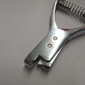 May include: A silver metal tool with a spring and two brass rivets. The tool is used for punching holes in leather or fabric.