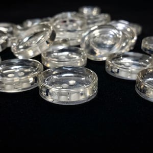 Peut inclure: Une collection de boutons ronds, clairs et transparents. Chaque bouton présente une surface lisse et brillante avec deux petits trous au centre pour la fixation. Les boutons sont dispersés, montrant leur forme circulaire.