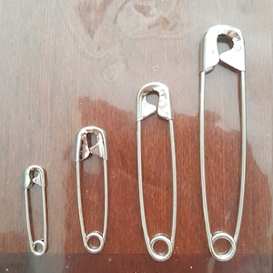 May include: Four silver safety pins of different sizes on a wooden surface.