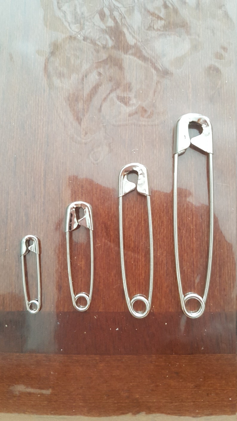 May include: Four silver safety pins of different sizes on a wooden surface.